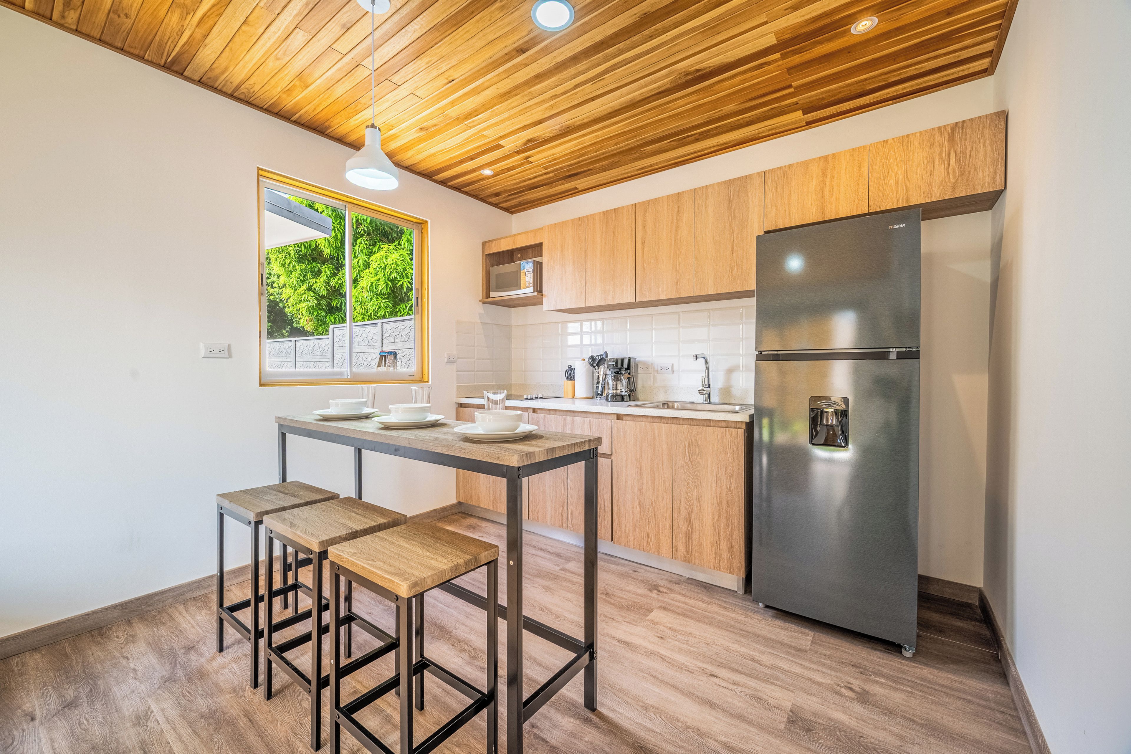 A modern kitchen with wooden cabinets, a stainless steel refrigerator, and a dining table with stools.