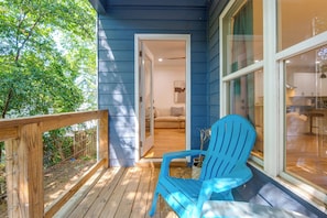 Eine Holzterrasse mit einem blauen Adirondack-Sessel, eine Glastür, die in einen Raum mit einer Couch und einer Lampe führt, und ein Blick auf eine Küche durch ein Fenster.