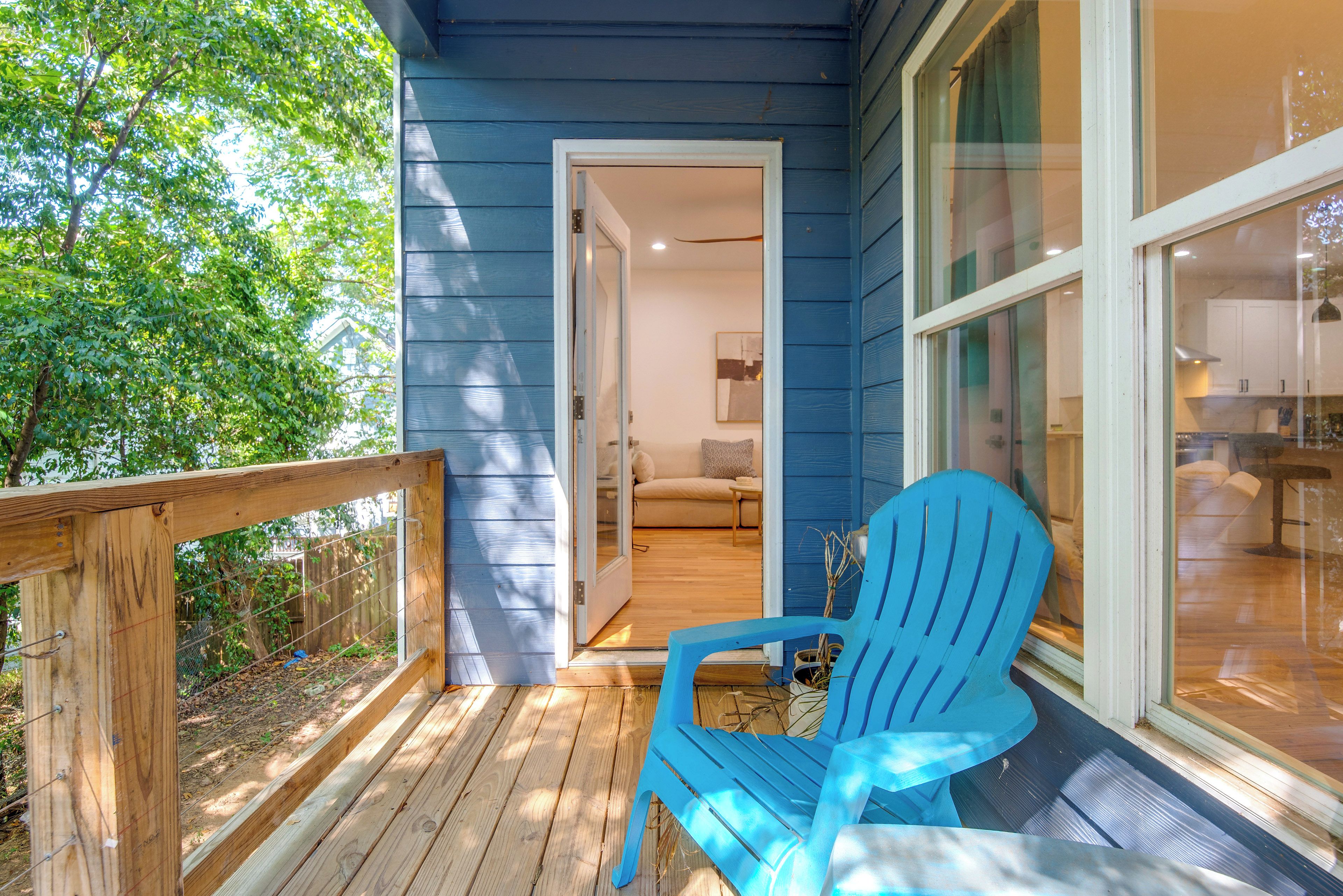 A wooden deck with a blue Adirondack chair, a glass door leading to a room with a sofa and a lamp, and a view of a kitchen through a window.