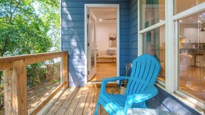 Una terraza de madera con una silla Adirondack azul, un ventanal que da a una habitación con sofá y lámpara, y vista a una cocina.