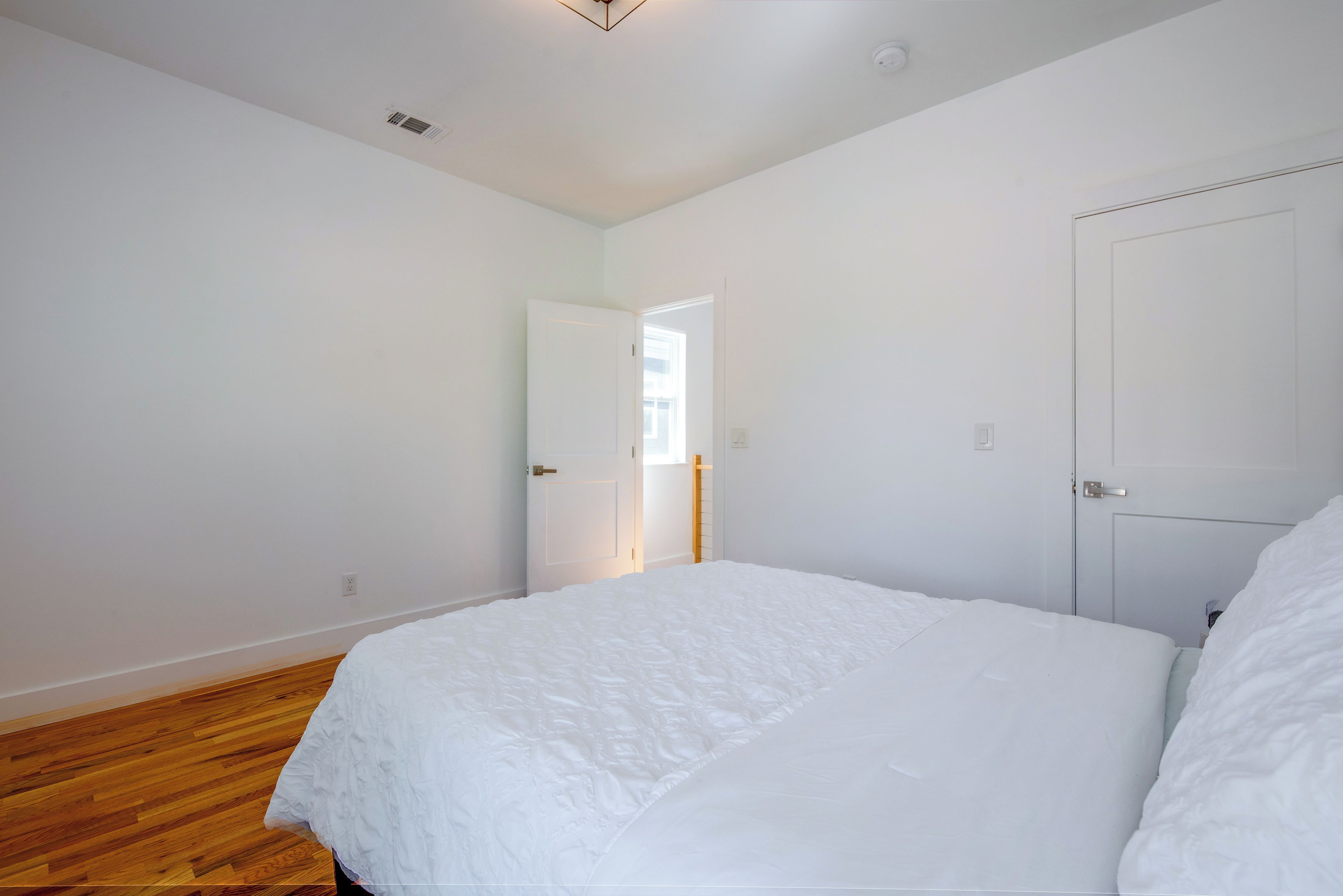 A bedroom with a large bed, wooden flooring, white walls, and a door leading to another room.