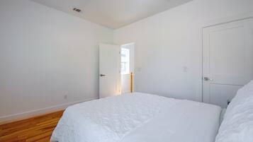 A bedroom with a large bed, wooden flooring, white walls, and a door leading to another room.