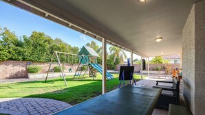 A covered patio area with a swing set, a slide, and a seating area with benches.