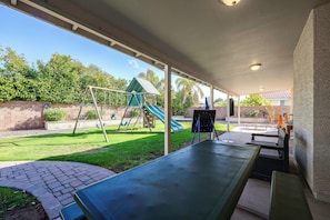 A covered patio area with a swing set, a slide, and a seating area with benches.