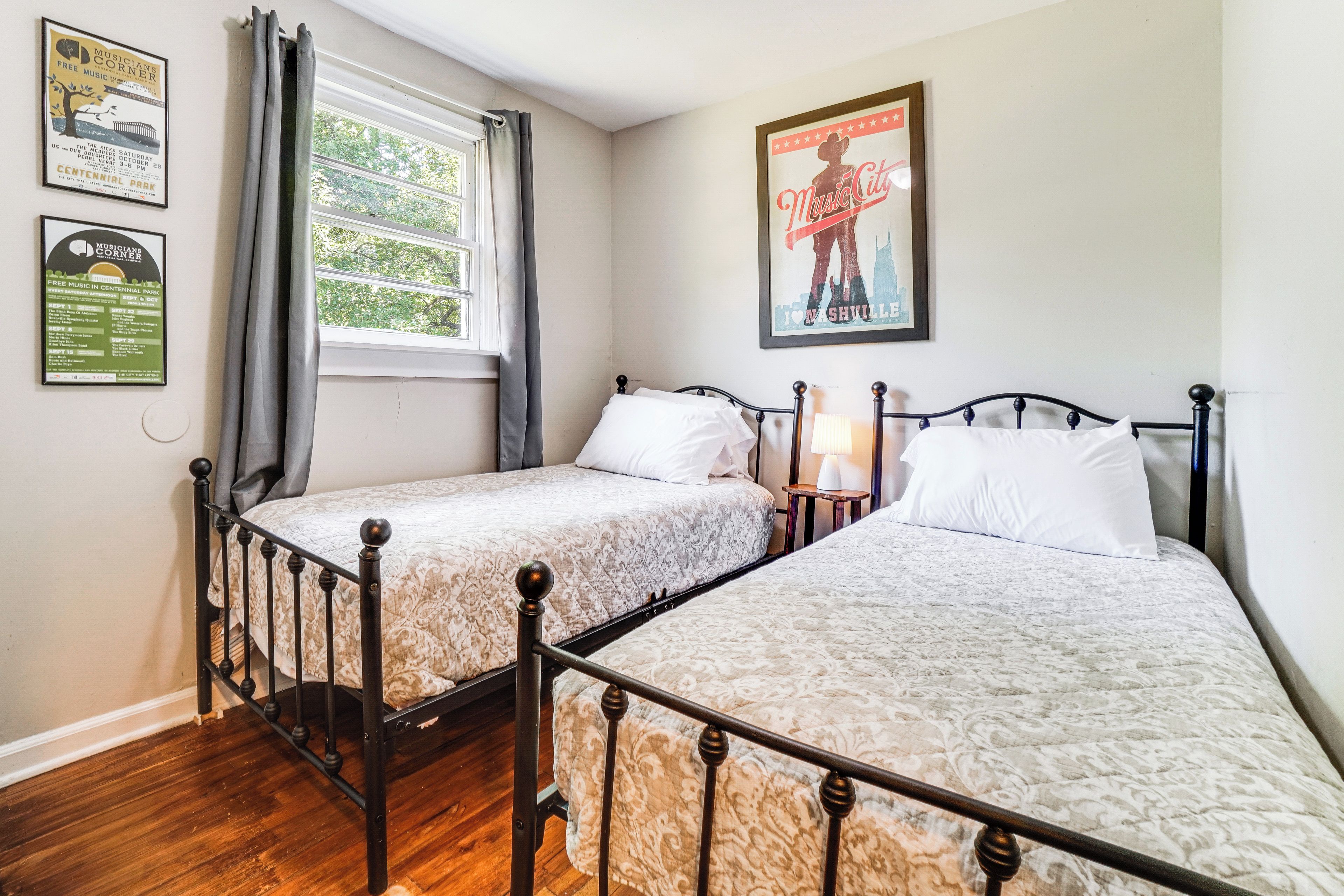 A hotel room with two single beds, a window with curtains, a poster on the wall, and a framed picture above the bed.