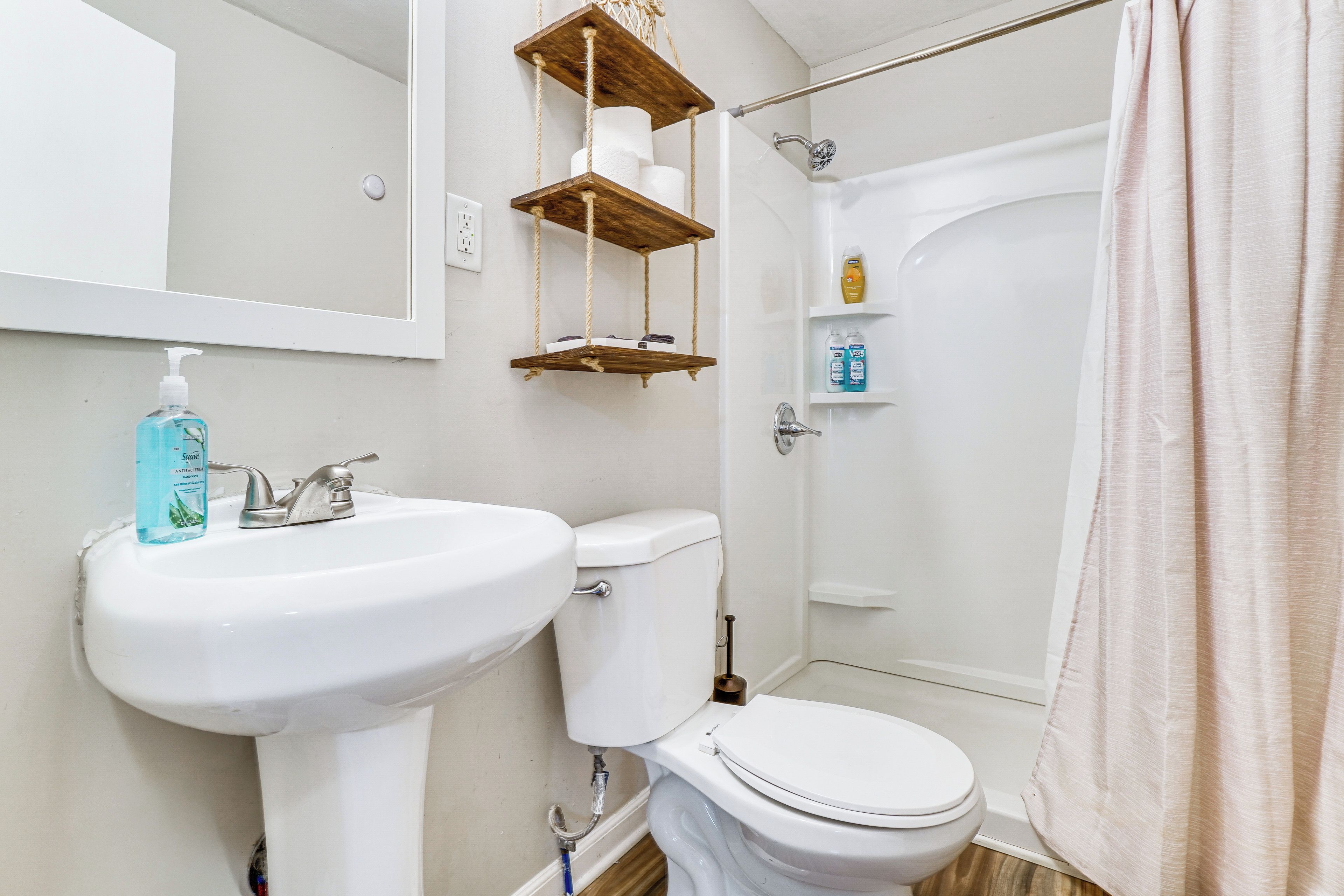 Un baño con lavamanos, inodoro y ducha blancos. Hay estantes de madera sobre el inodoro y una botella de jabón para manos en el lavamanos.