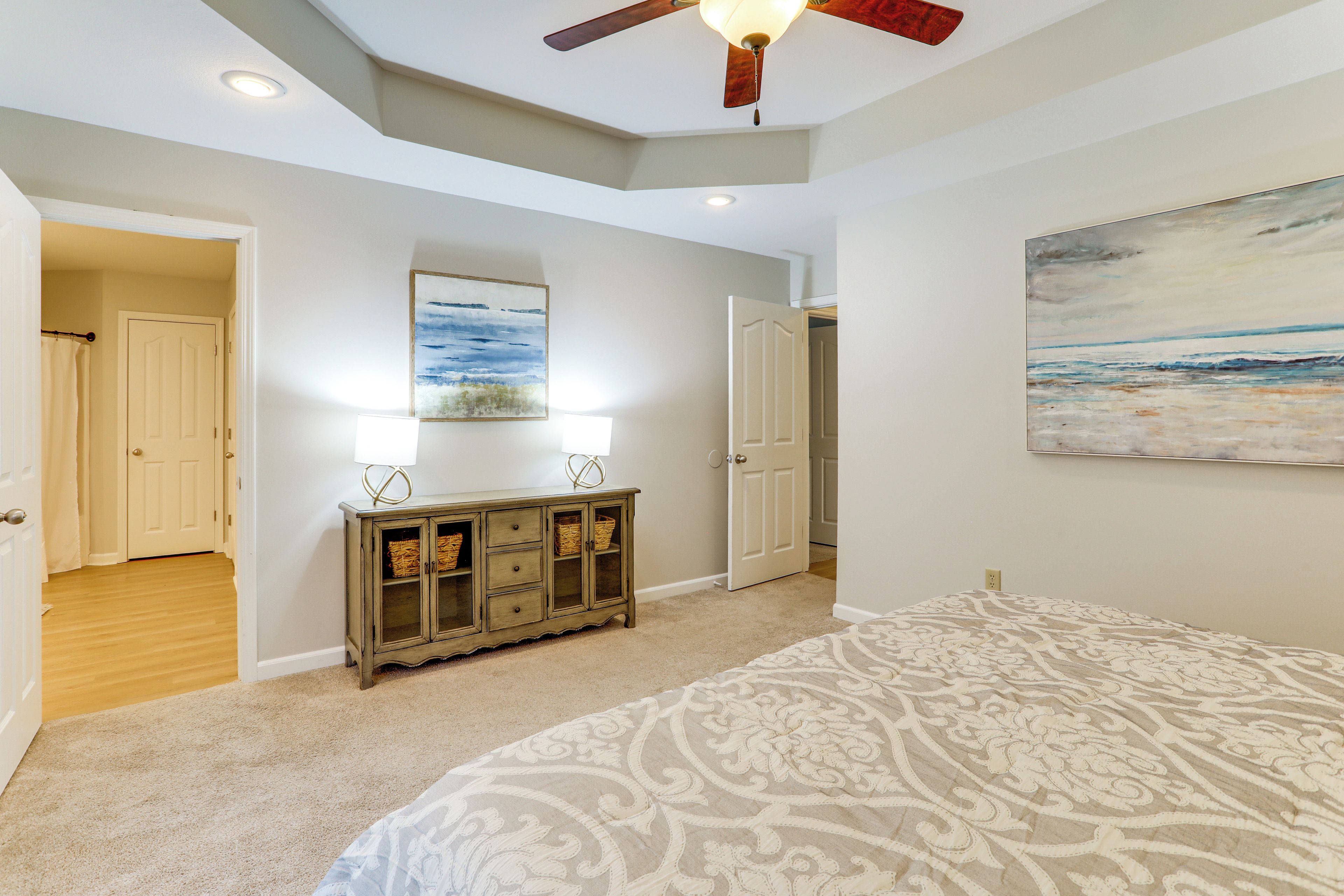 A bedroom with a ceiling fan, a bed with a patterned bedspread, a dresser with lamps, and a painting on the wall.