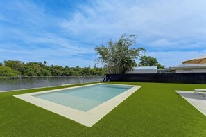 Piscina retangular cercada por grama sintética, com uma vedação e árvores ao fundo.