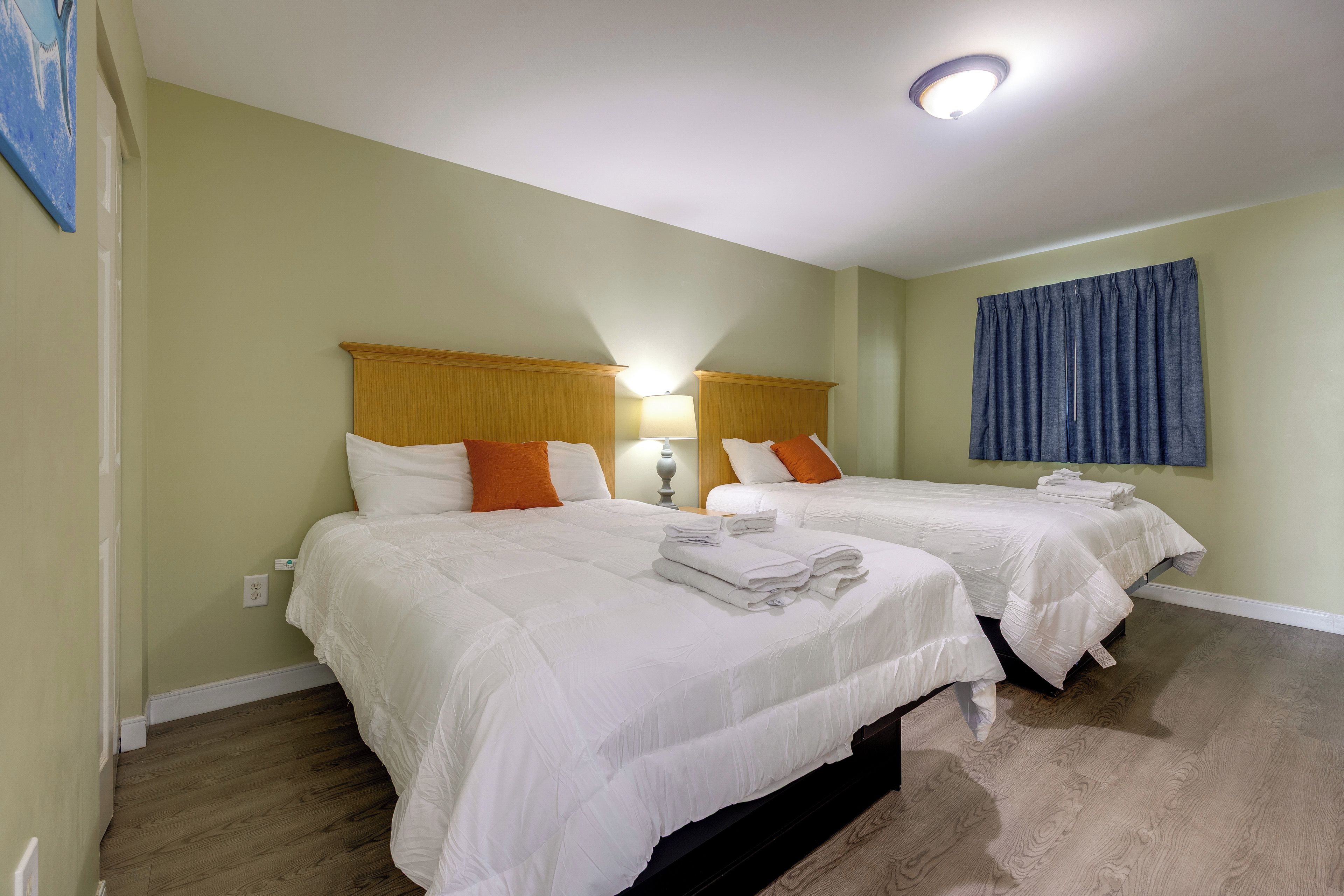 Quarto de hotel com duas camas, mesa de cabeceira, abajur e cortina azul.