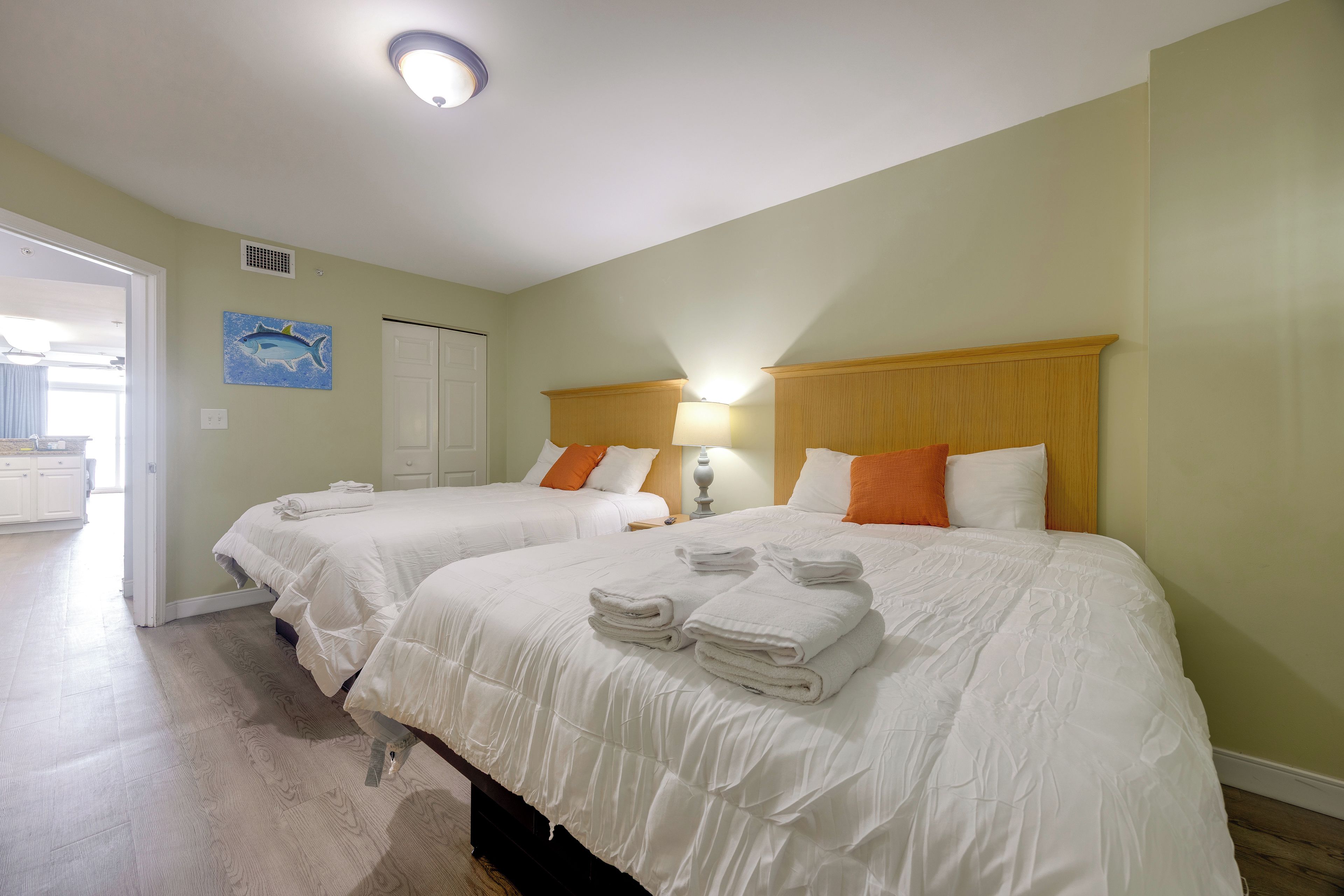 A hotel room with two beds, each with white bedding and folded towels. There is a door leading to a bathroom, and a wall-mounted light fixture above the beds.
