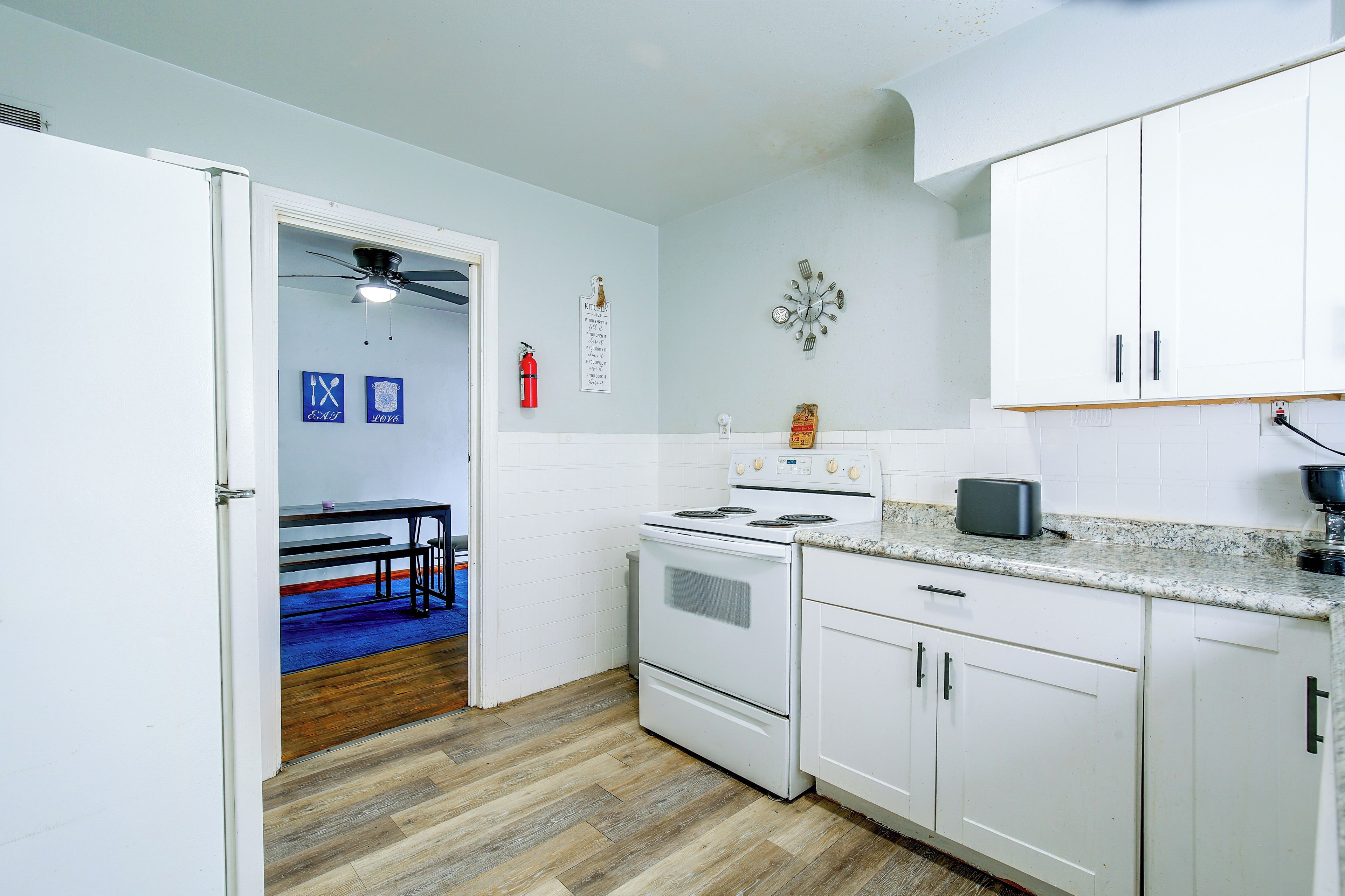 Una cocina con armarios blancos, una estufa y un microondas. Hay un tapete azul en el suelo y un ventilador de techo.