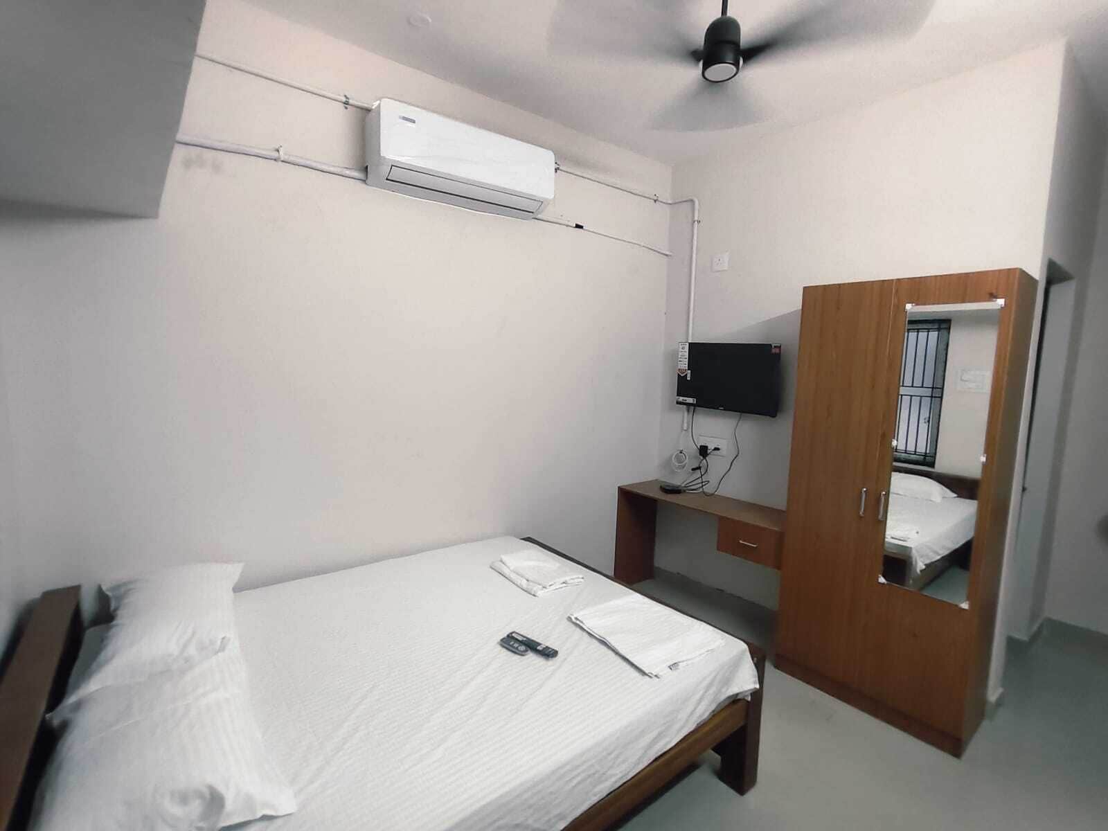 A small, single-bed room with a wooden desk, a flat-screen TV mounted on the wall, and a ceiling fan.