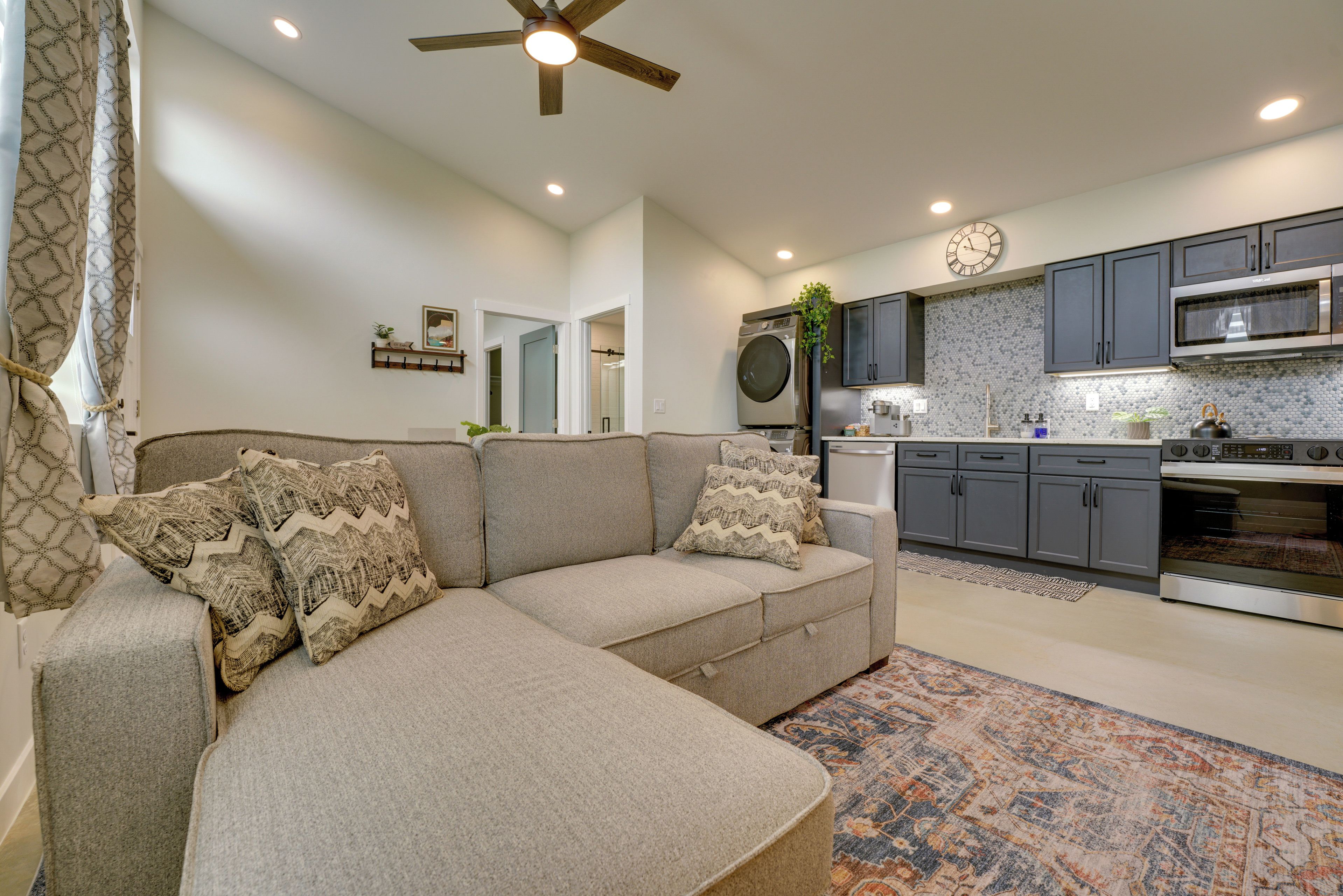 A modern living room with a sectional sofa, a kitchen with grey cabinets, and a ceiling fan.