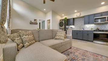 A modern living room with a sectional sofa, a kitchen with grey cabinets, and a ceiling fan.