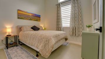 A bedroom with a bed, bedside lamp, a painting on the wall, a window with curtains, and a small cabinet.