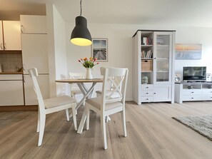 A modern kitchen with white cabinets, a dining table with chairs, a pendant light, and a bookshelf with books and decorative items. - Holiday Apartment With 2 Bedrooms (Varel)