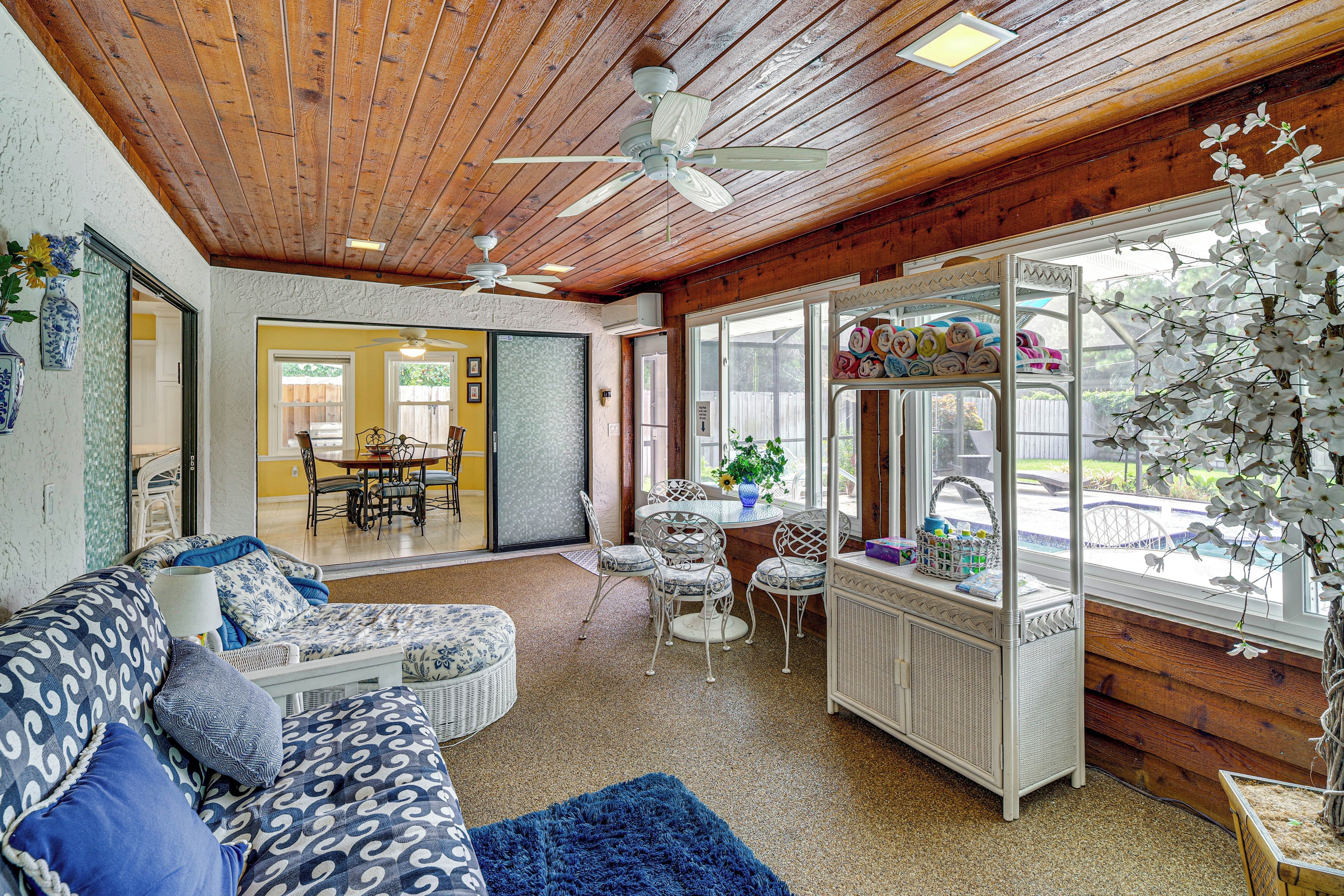 A cozy room with a wooden ceiling, a ceiling fan, a dining area with a table and chairs, and a large window with white flowers.