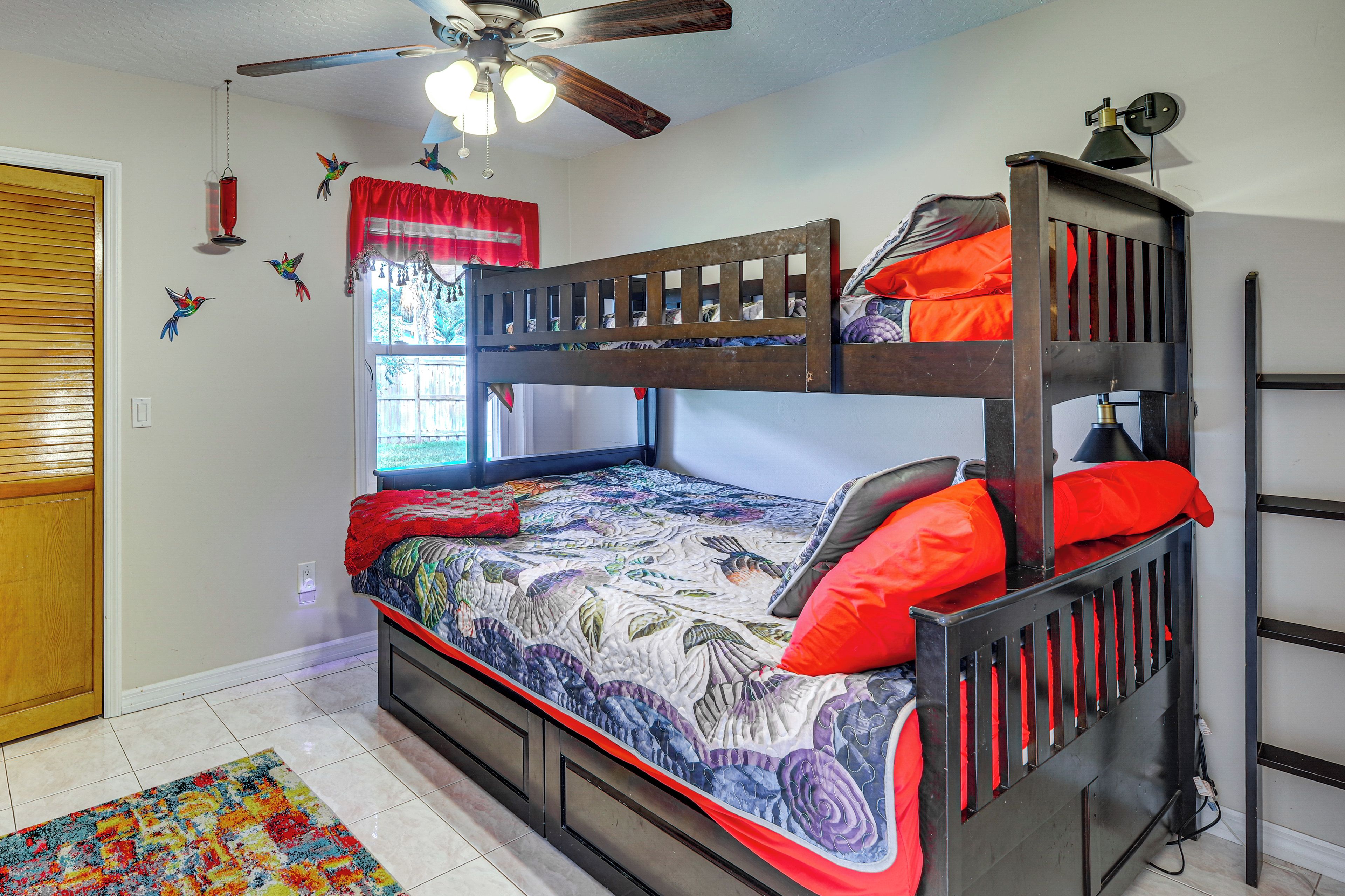 A bunk bed with a colorful quilt, a ceiling fan, and a window with red curtains.