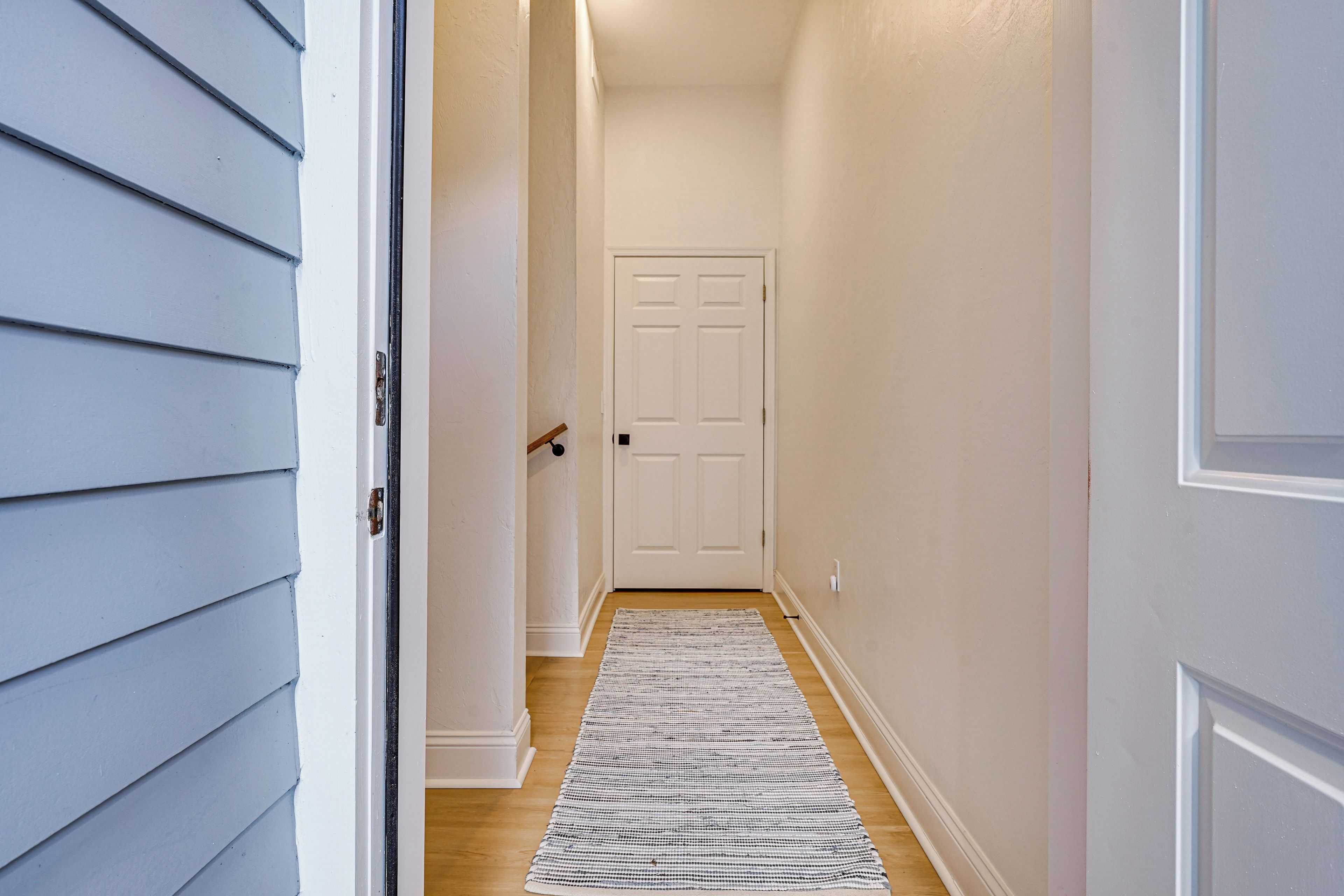Un couloir avec une porte blanche au fond, un tapis à motifs sur le sol et un interrupteur sur le mur.