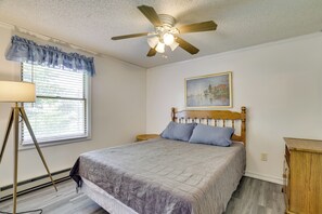 A bedroom with a bed, a ceiling fan, a window with blinds, a bedside lamp, and a painting on the wall.