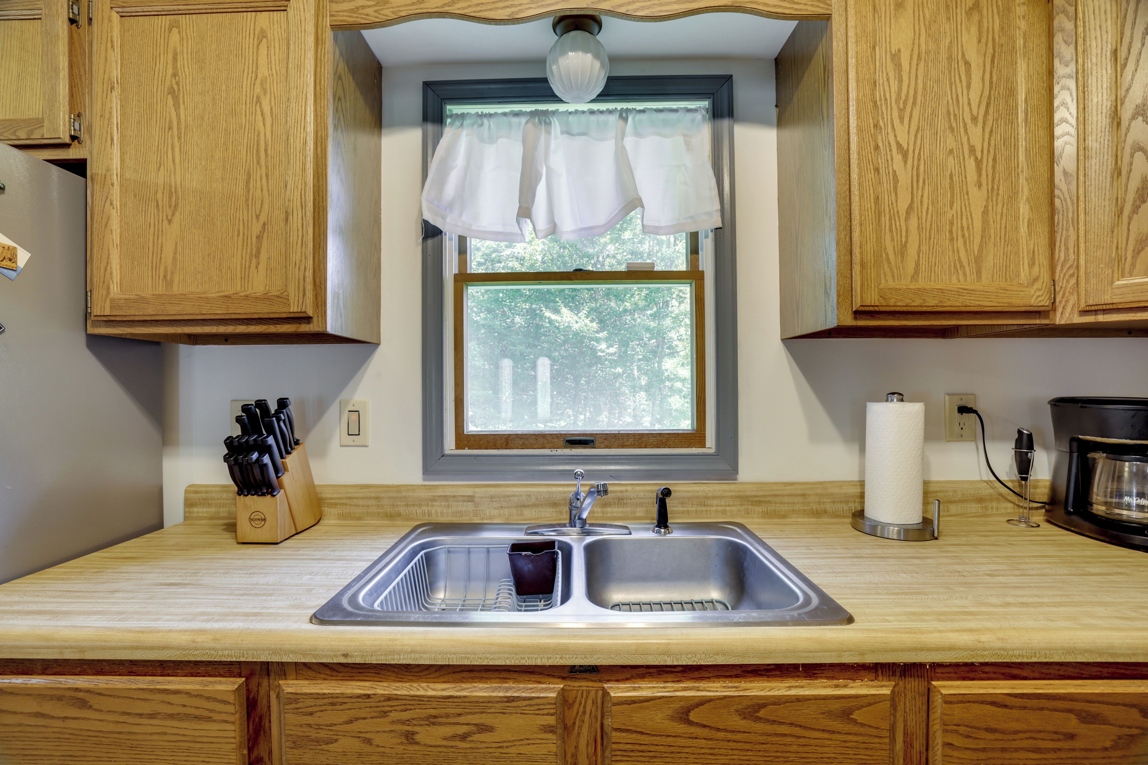 A kitchen with wooden cabinets, a double sink, a window with a view of trees, and a coffee maker.