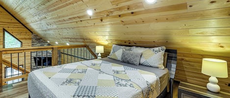 Un dormitorio con techo de madera, una cama con colcha estampada, dos lámparas de noche y una ventana con vistas a la vegetación.