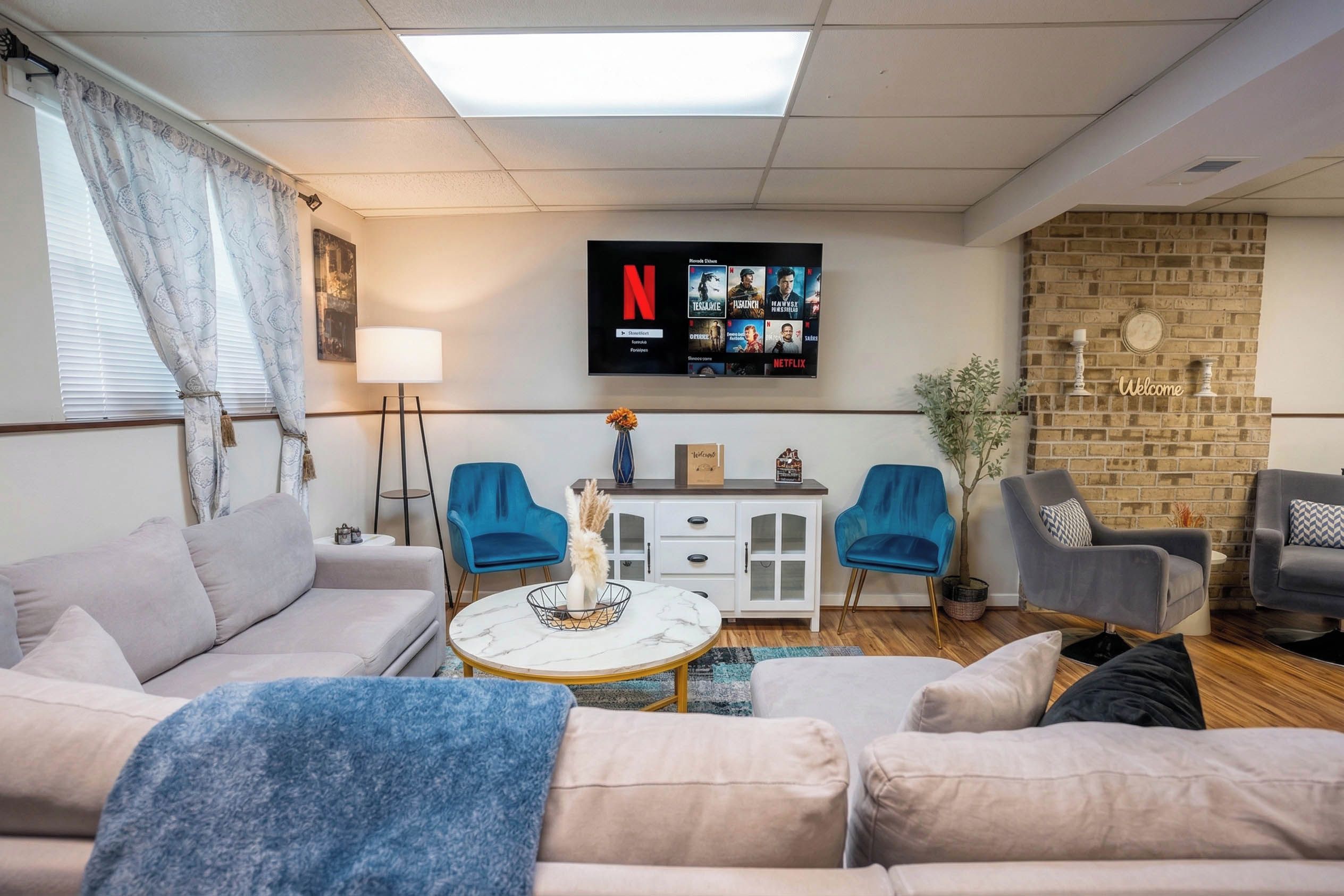 A living room with a sofa, two armchairs, a coffee table, a TV mounted on the wall, and a brick wall.