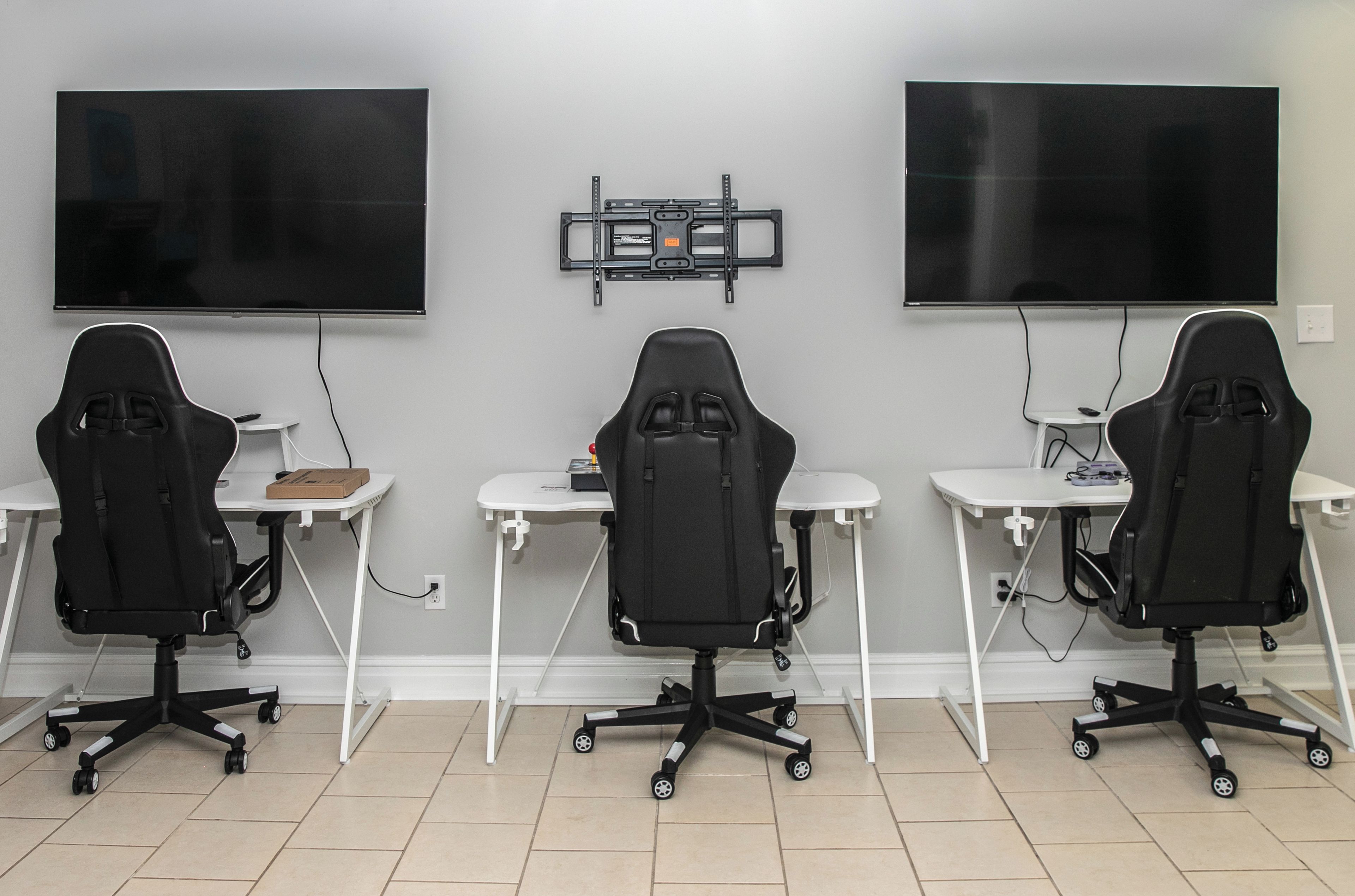 A room with three gaming chairs, two desks, and two large flat-screen TVs mounted on the wall.
