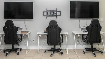 A room with three gaming chairs, two desks, and two large flat-screen TVs mounted on the wall.