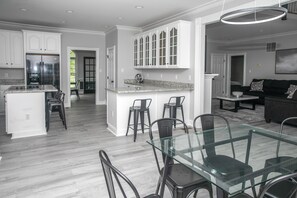 A modern kitchen with a central island, bar stools, and a dining area with a glass table and chairs. - Waterfront Deck, Family Arcade! Lake Gaston Home (Bracey)