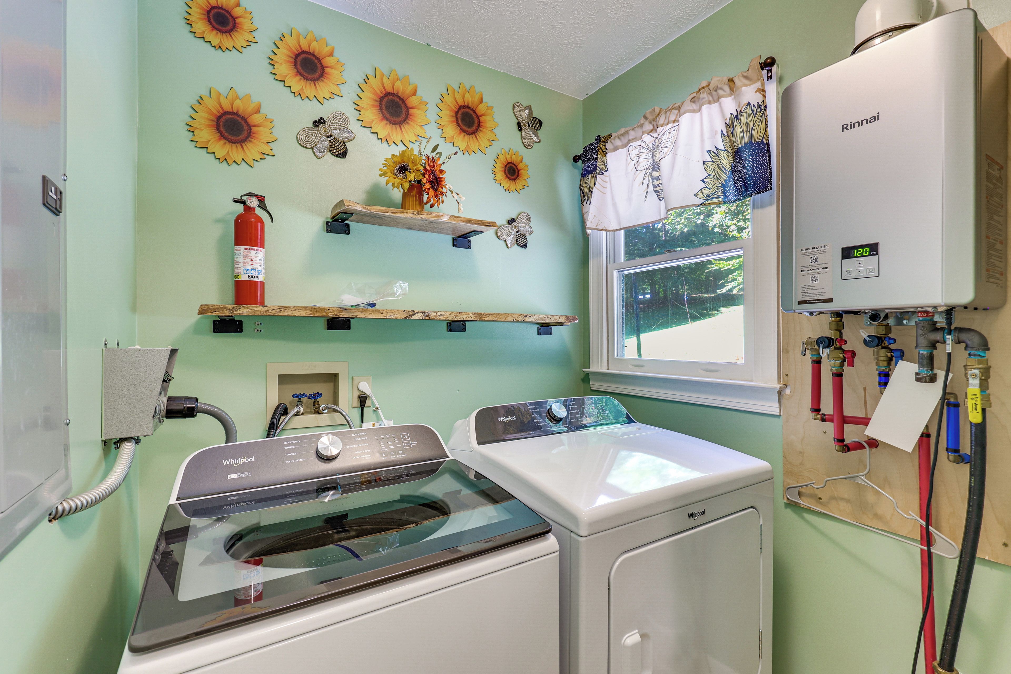 A laundry room with a Whirlpool washer and dryer, a fire extinguisher, and a window with a view of greenery.