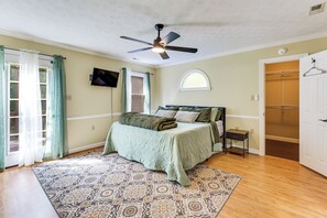 A bedroom with a bed, a ceiling fan, a TV, a window with curtains, a patterned rug, and a door with a coat rack.