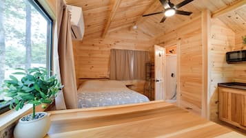 A wooden cabin interior with a bed, a ceiling fan, a window with curtains, and a potted plant.