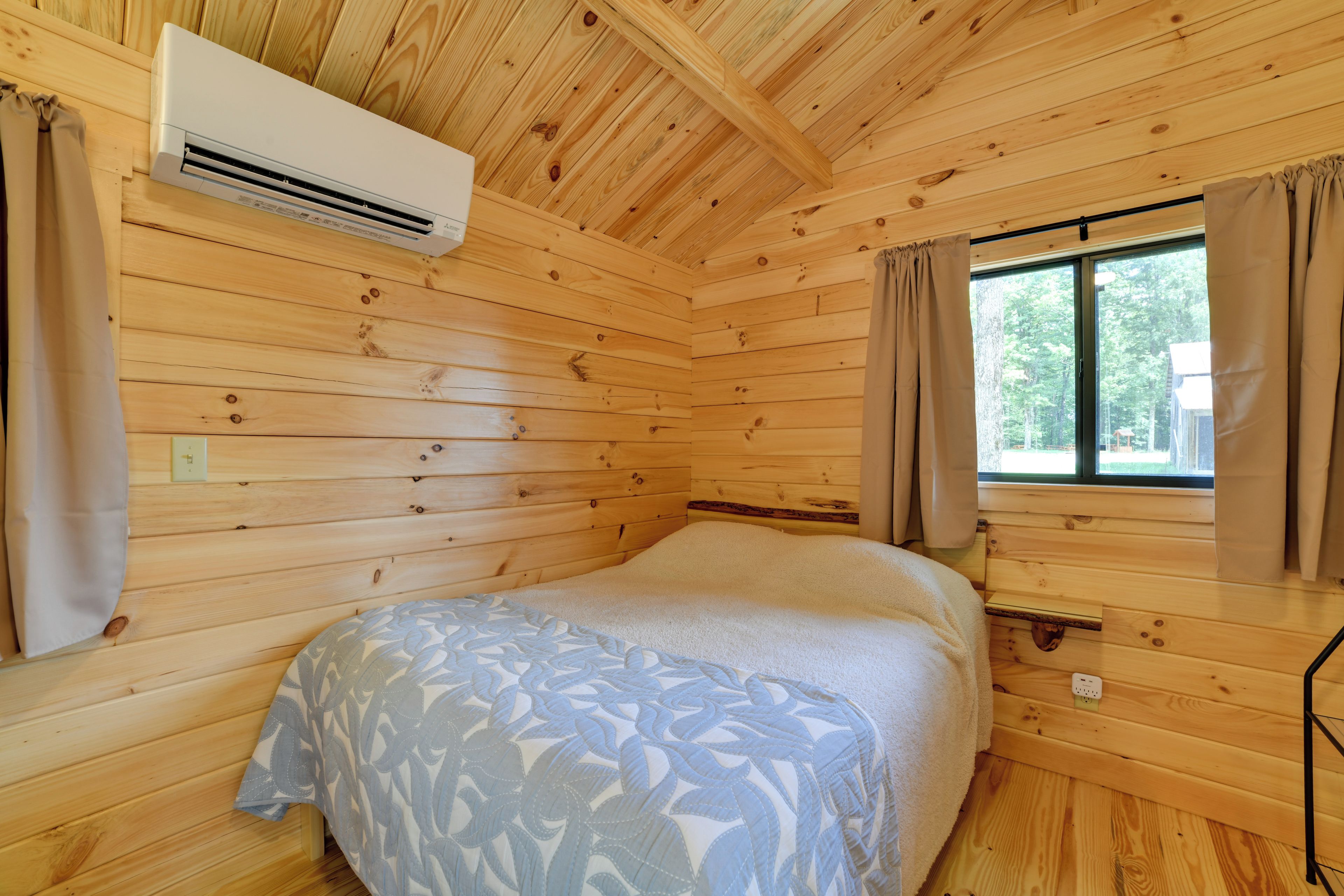 A wooden cabin room with a bed, an air conditioner, a window with curtains, and a power outlet.