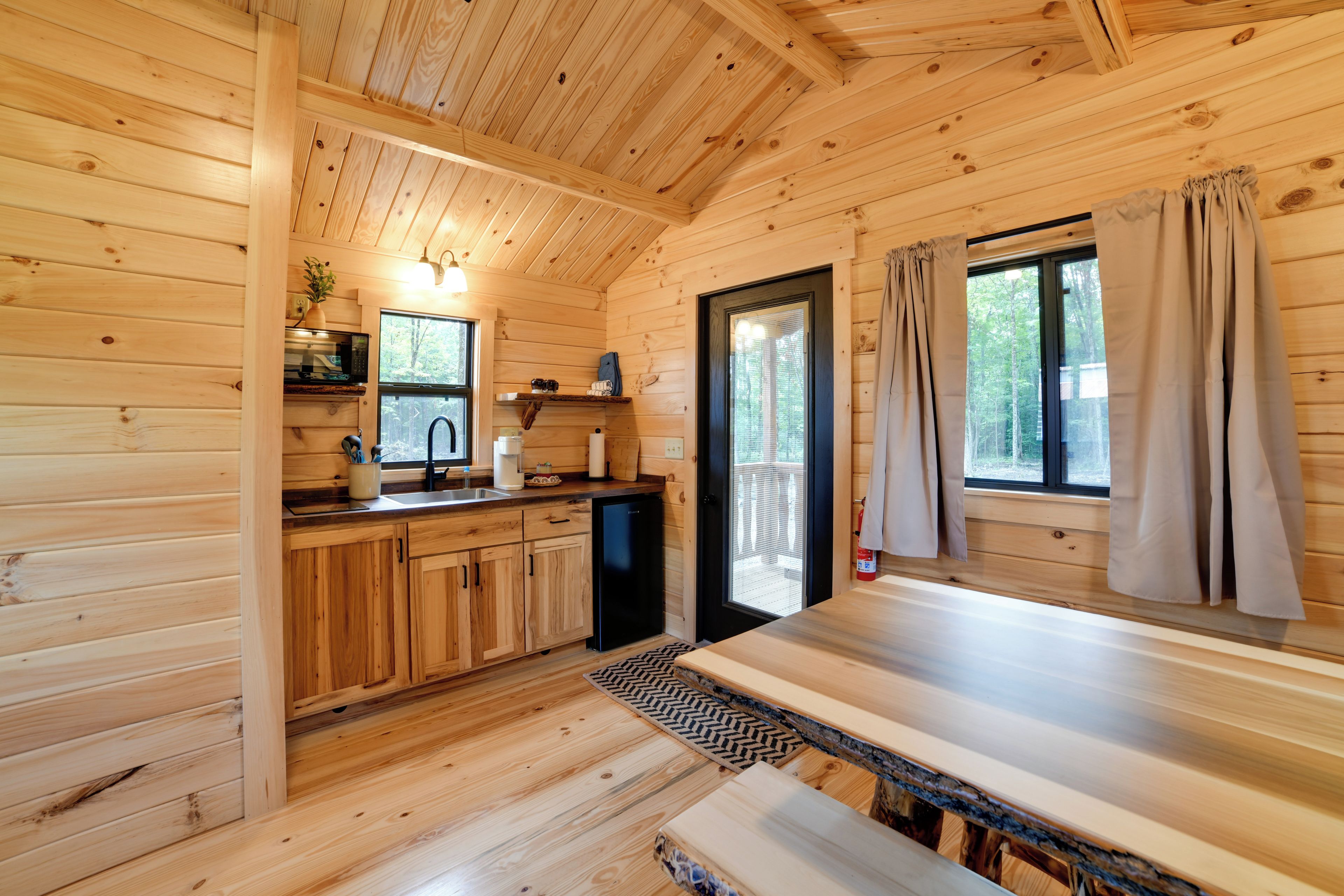 A wooden cabin interior with a kitchen, a dining area, and a window with curtains.