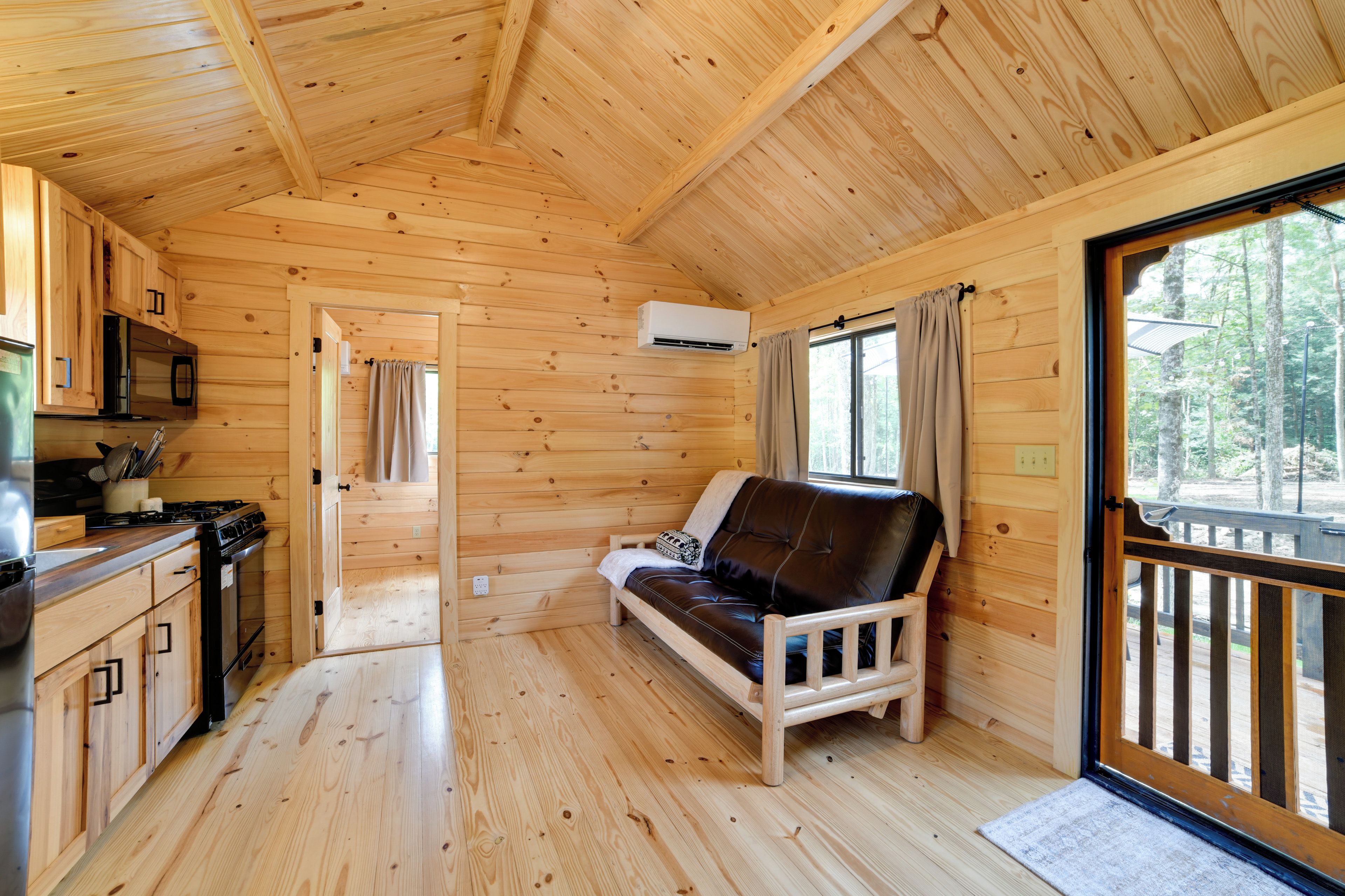 A wooden cabin interior with a kitchenette, a sofa, and a window overlooking a forest.