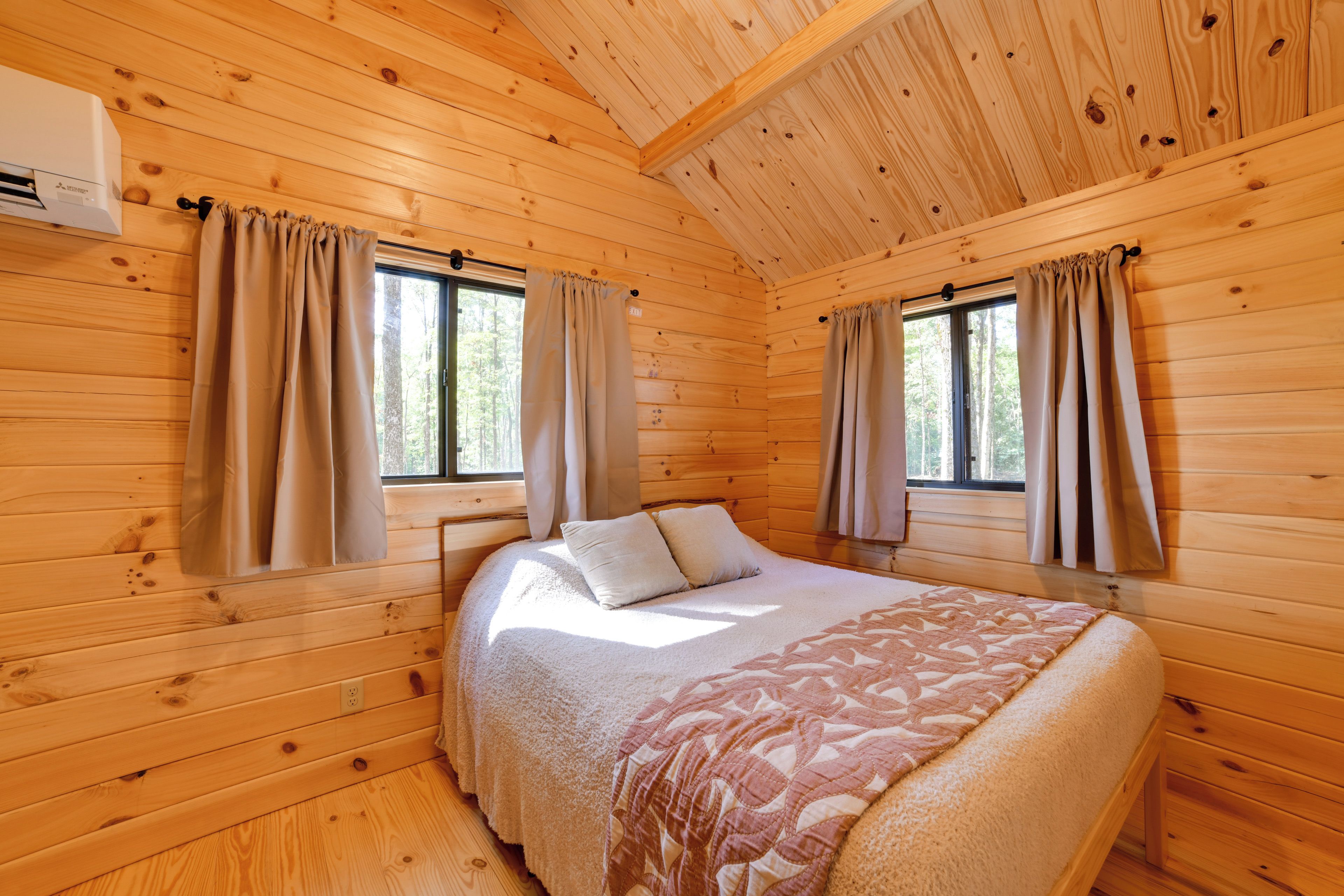 A wooden cabin room with a bed, two windows with curtains, and an air conditioning unit.
