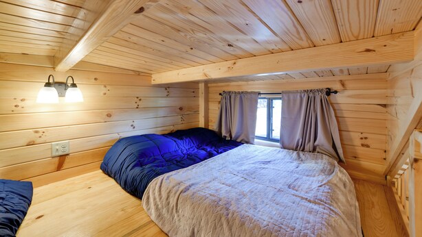 Ett sovrum i en stuga med träpanel, en säng, två sänglampor och ett fönster med gardiner.