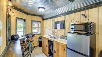 A compact kitchen with a stainless steel refrigerator, a microwave, and a sink. There are wooden walls and a window with an American flag.