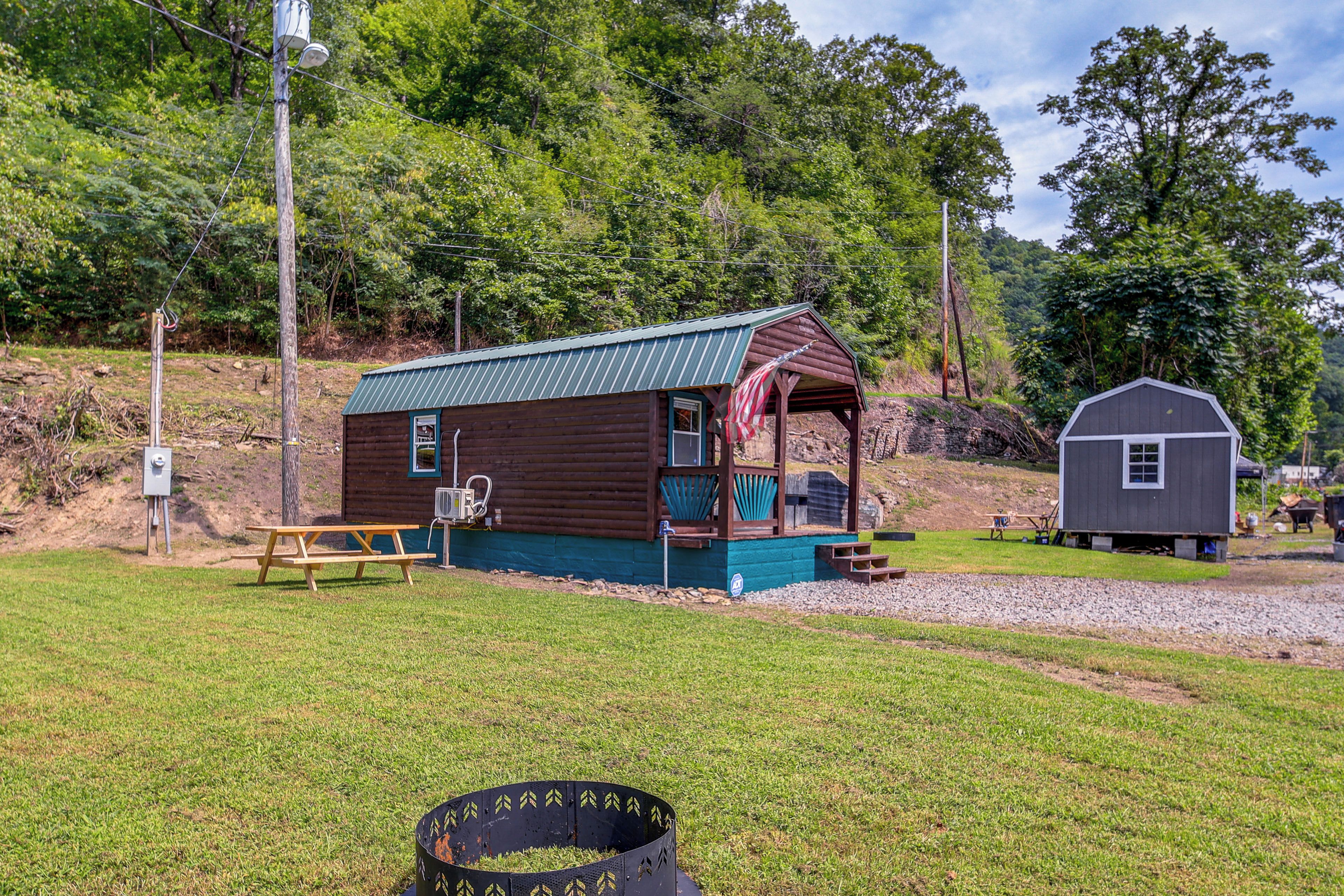 A small wooden cabin with a porch, a picnic table, and a shed in a grassy area with trees in the background.