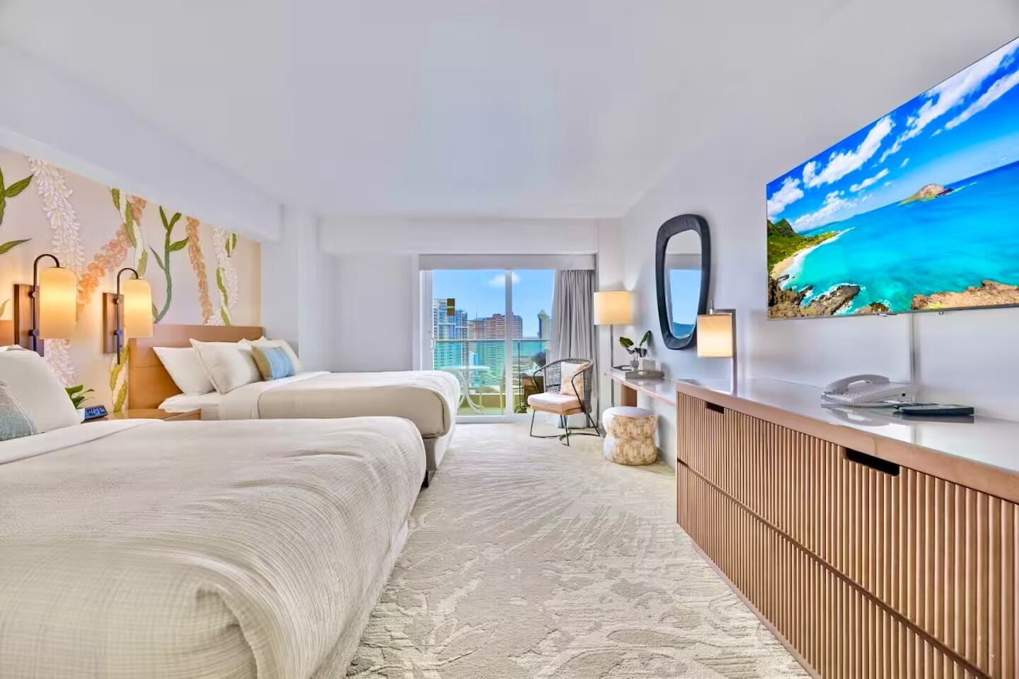 A hotel room with two beds, a large window with a city view, a desk with a flat-screen TV, and a decorative wall with a floral pattern.