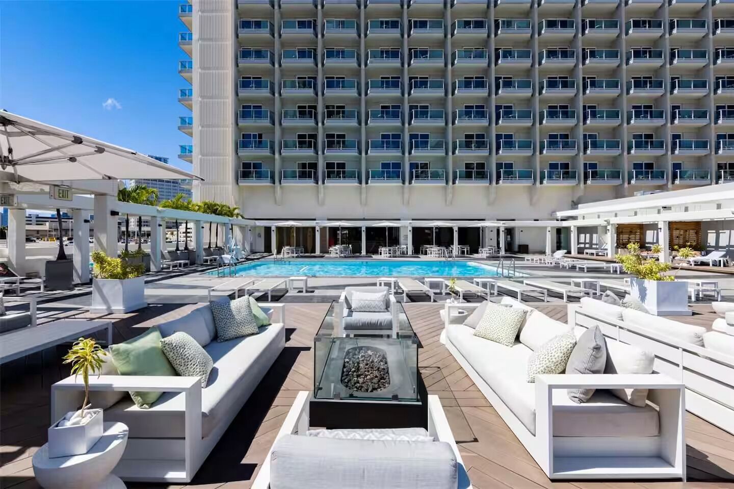 A rooftop pool area with lounge chairs and sofas, a large pool, and a high-rise building in the background.