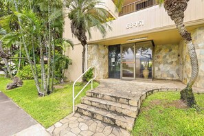 A building entrance with a stone staircase, palm trees, and a glass door with a number plaque.