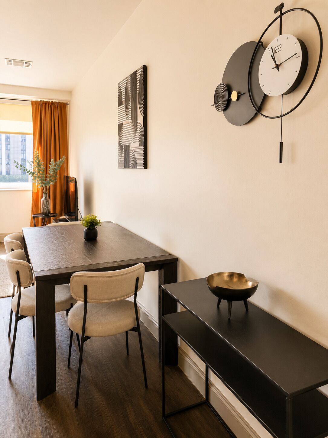 Un comedor con una mesa de madera, sillas y un banco negro. Hay un reloj de pared y una obra de arte enmarcada sobre la mesa.