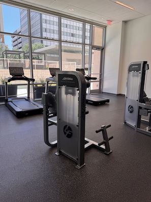 A gym with cardio equipment and weight machines, large windows, and a view of buildings outside.