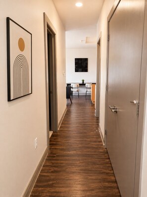 A hallway with wooden flooring, a framed artwork on the left, and a kitchen area in the background.