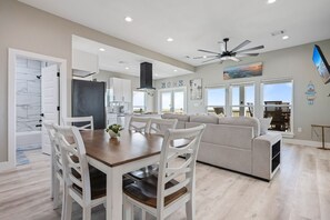 A modern kitchen with white cabinetry, a dining area with a wooden table and white chairs, and a grey sofa in a living area with a cityscape view. - The Blue Crab - On Canal, boat slip w/lift, Bay access, outdoor bar (Gilchrist)