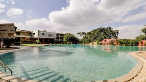 A clear pool with a surrounding deck, a modern building with multiple floors, and lush greenery in the background. - Gardeenia Comfortes  (Bommasandra)