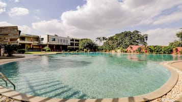 Una piscina cristalina con un deck circundante, un edificio moderno de varios pisos y abundante vegetación al fondo.