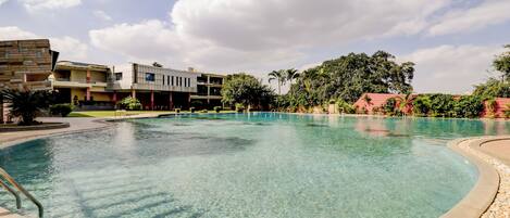A clear pool with a surrounding deck, a modern building with multiple floors, and lush greenery in the background.